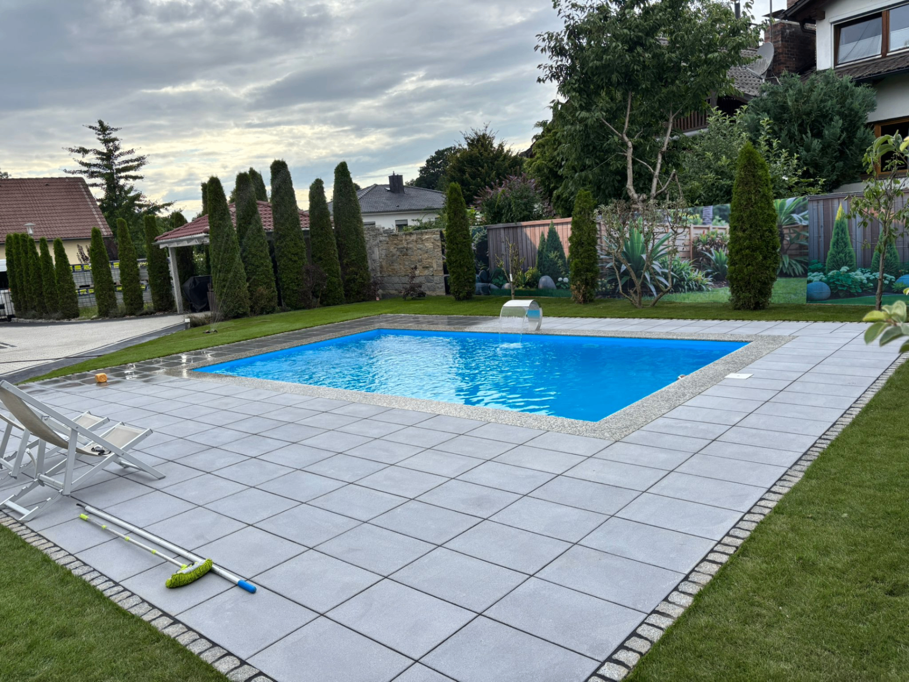 Privater Garten mit rechteckigem Pool, großflächiger Terrasse aus grauen Betonplatten, angrenzendem Rasen und Thuja-Hecke im Hintergrund